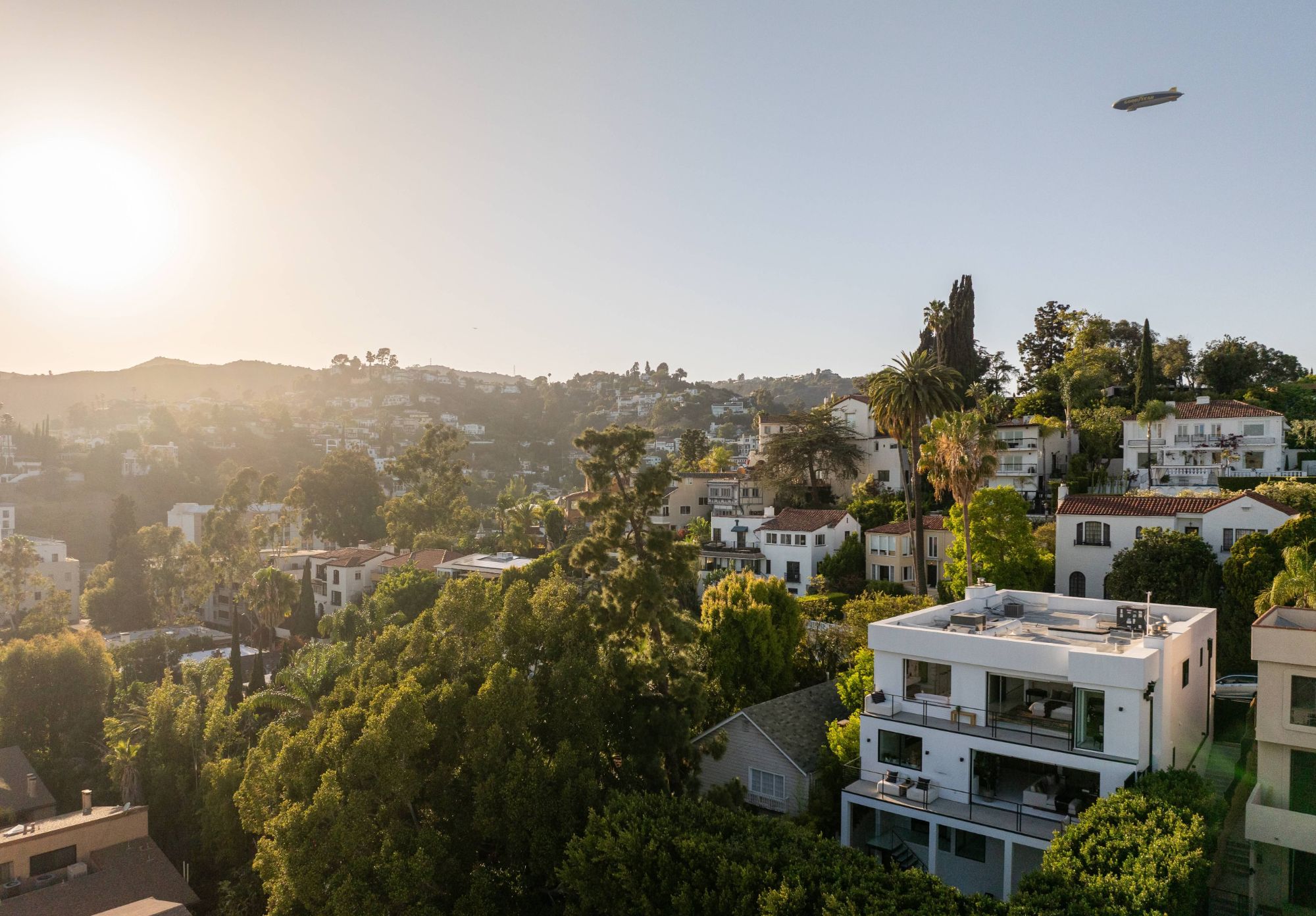The house among the hillside homes of Whitley Heights at golden hour, with the Hollywood Hills behind and the Goodyear blimp overhead